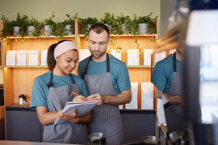 Waist up portrait of two young waiters wearing aprons and holding clipboard while doing inventory in cafe or coffee shop, copy spaceの写真素材