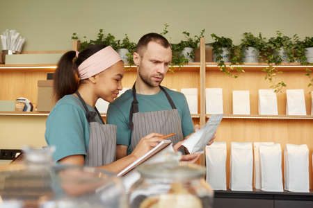 Waist up portrait of two young waiters wearing aprons while doing inventory in cafe or coffee shop,...