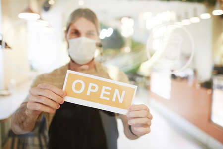 Close up of male cafe worker wearing mask and holding OPEN sign on glass door, copy spaceの写真素材