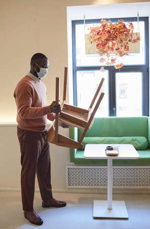 Full length portrait of African-American man wearing mask in cafe and arranging furniture while preparing for opening in morningの写真素材
