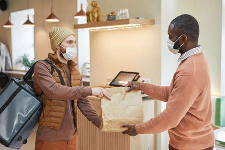 Side view portrait of African-American man handing order to delivery man in cafe, both wearing...