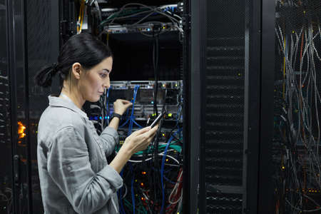 Side view portrait of female network technician connecting cables in server cabinet while setting up...