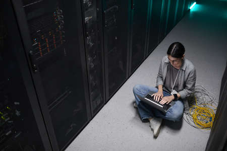 High angle view at female data engineer using laptop while sitting on floor in server room and setting up supercomputer network, copy spaceの写真素材