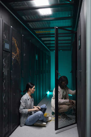 Vertical portrait of two female data engineers using laptop in server room and setting up...