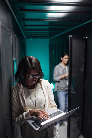 Vertical portrait of female data engineer using laptop in server room while setting up supercomputer...