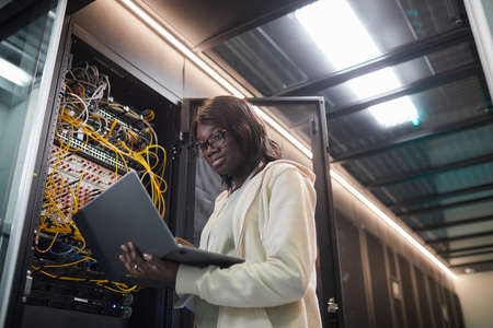 Low angle portrait of African-American female network engineer standing by server cabinet and holding laptop while working with supercomputer in data center, copy spaceの写真素材