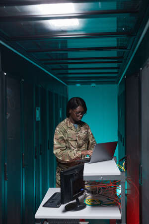 Vertical portrait of young African-American woman wearing military uniform using computer while setting up network in server roomの写真素材
