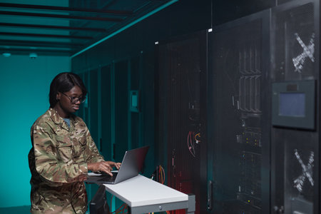 Waist up portrait of young African-American woman wearing military uniform using computer while setting up network in server room, copy spaceの写真素材
