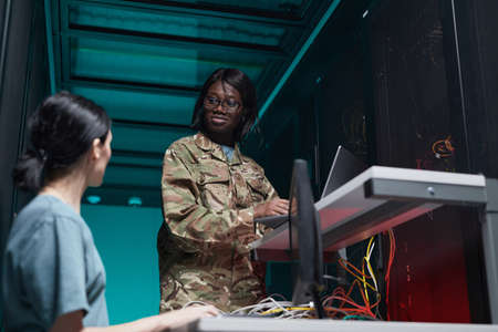 Low angle portrait of young African-American woman wearing military uniform using computer while setting up network in server roomの写真素材