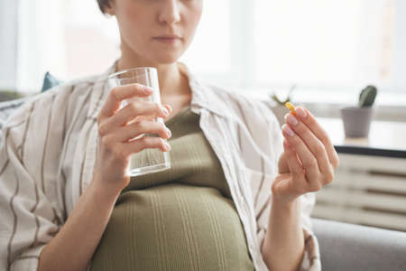 Close-up of pregnant woman holding glass with water and drinking vitamins in the morningの写真素材