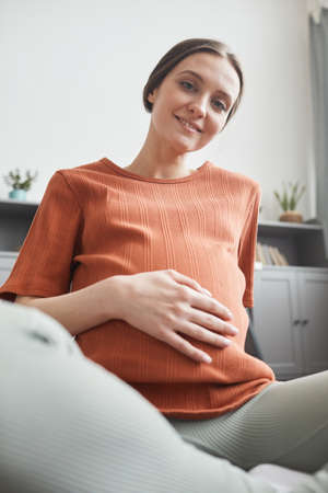 Portrait of happy pregnant woman sitting on sofa at home and smiling at cameraの写真素材