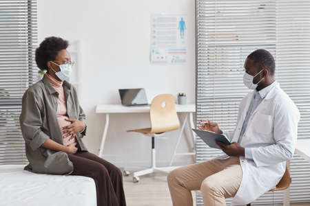 Pregnant woman in protective mask visiting the doctor at hospital and talking to himの写真素材