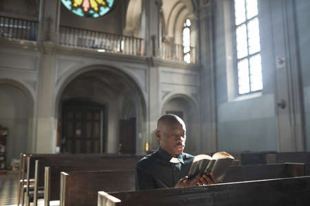 African priest in collar sitting on the bench in church and reading the Bibleの写真素材