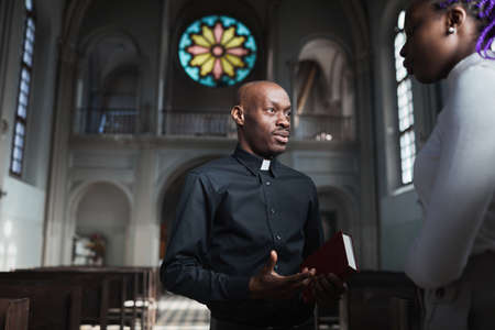 African young priest talking to woman and discussing the parts of Bible standing in the churchの写真素材