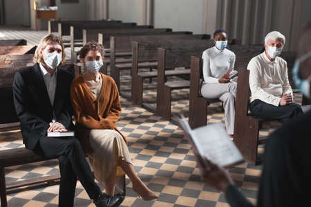Group of people in protective masks sitting on bench and listening to mass in the church during pandemicの写真素材