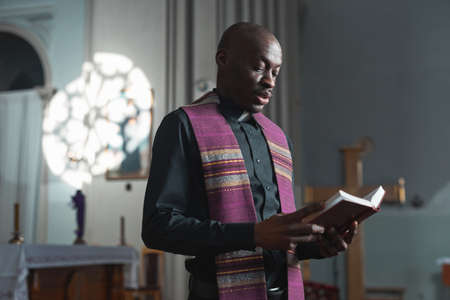 African young priest reading the Bible during mass while standing near the altar in churchの写真素材
