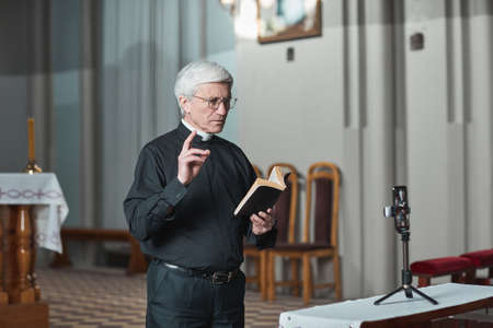 Senior priest reading Bible and shooting his performance on mobile phone while standing in the churchの写真素材