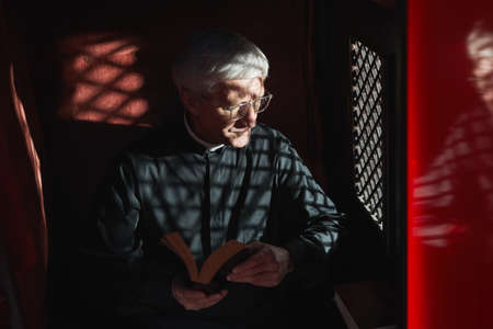 Senior priest sitting with the Bible in confessional and listening to confessionの写真素材