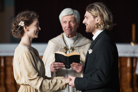 Young couple looking at each other and smiling during wedding ceremony with priest in the churchの写真素材