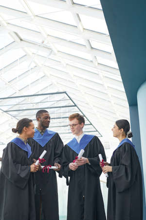 Vertical view at diverse group of young people wearing graduation gowns indoors in modern university...