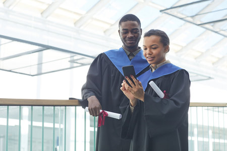 Waist up portrait of young African-American couple wearing graduation gowns while taking selfie indoors in modern university interior, copy spaceの写真素材