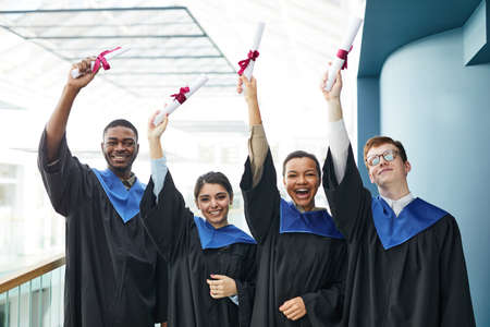 Waist up view at diverse group of cheerful young people wearing graduation gowns and smiling at...