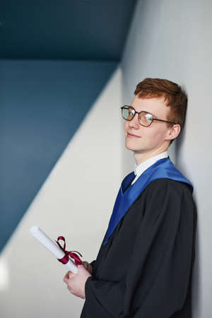 Graphic vertical portrait of young man wearing graduation gown and holding diploma while smiling at camera in collegeの写真素材