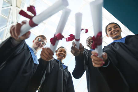 Low angle view at group of university graduates holding diplomas to camera while standing indoorsの写真素材