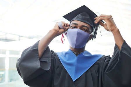 Waist up portrait of young African-American man wearing mask and graduation gown while smiling happily at camera indoors, copy spaceの写真素材