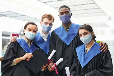 Diverse group of young people wearing graduation gowns and masks while looking at camera indoors in college interiorの写真素材