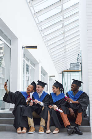 Full length view at diverse group of happy young people wearing graduation gowns and taking selfie while sitting on steps outdoorsの写真素材