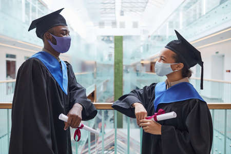 Side view portrait of two African-American young people wearing graduation gowns and masks while chatting indoorsの写真素材