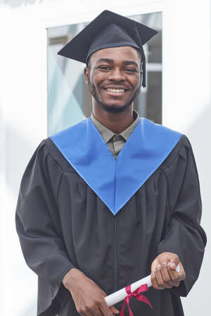 Vertical portrait of African-American young man wearing graduation gown and hat smiling at camera happilyの写真素材