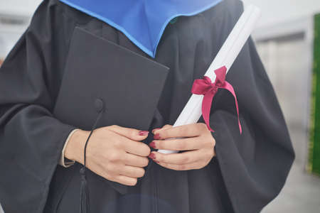Cropped portrait of unrecognizable young woman holding diploma and graduation cap while standing...