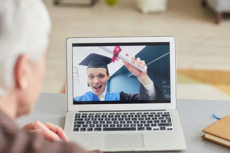 Closeup of mature man watching teenage son graduating by video chat on laptop screen