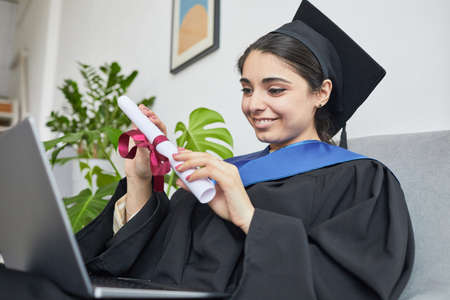Portrait of young woman wearing graduation gown and using laptop while speaking by video chat...