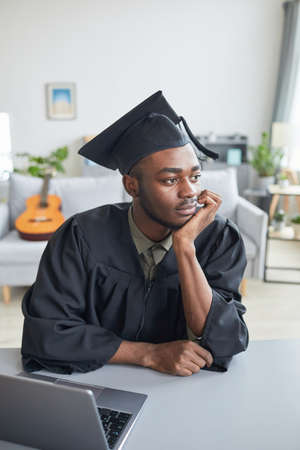 Vertical portrait of pensicve African-American man wearing graduation gown at homeの写真素材