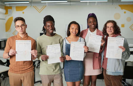 Diverse group of young people standing in school classroom smiling happily at camera and holding test certificates, copy spaceの写真素材