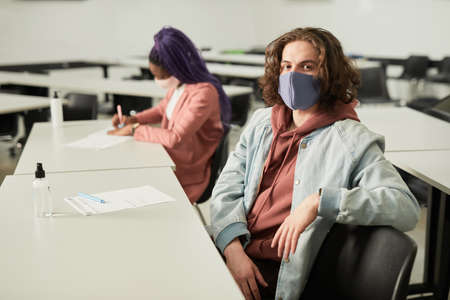Portrait of long haired young man wearing mask in school and looking at camera while sitting at desk in classroom, copy spaceの写真素材