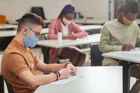 Side view portrait of young man wearing mask while taking test or exam in school with diverse group of people, copy spaceの写真素材