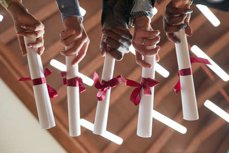 Low angle closeup of young people holding diplomas with red ribbon during graduation ceremony in college, copy spaceの写真素材