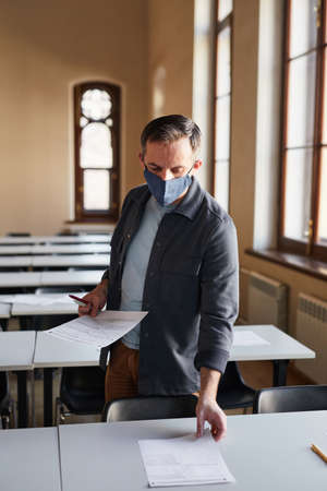 Vertical portrait of mature college professor wearing mask while laying out exam papers in school auditorium lit by sunlightの写真素材
