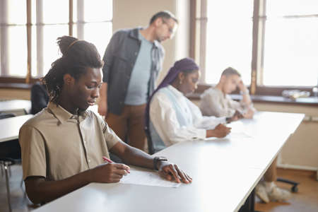 Diverse group of students taking exam in college while sitting in row at desk in auditorium, focus on young African-American man in foreground, copy spaceの写真素材