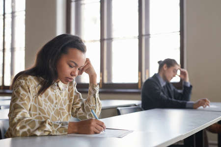 Side view portrait of two students taking exam in row while sitting at desk with social distancing, focus on young African-American woman in foreground, copy spaceの写真素材