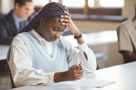Portrait of young African-American woman taking exam in school while sitting at desk in college and...