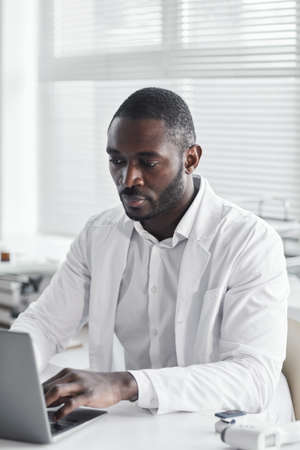 African young doctor in white coat sitting at the desk and working on laptop at his officeの写真素材