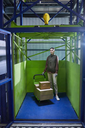 Young woman standing with trolley with boxes and looking at camera while working in the warehouse