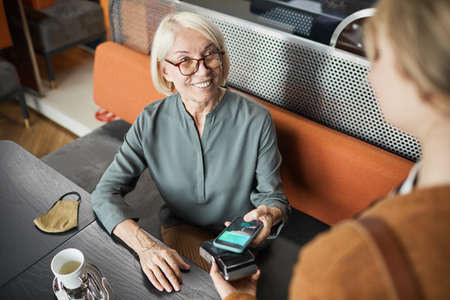 Positive mature cafe guest in eyeglasses sitting at table with cloth mask and paying for lunch with...
