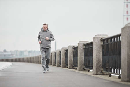 Purposeful senior Caucasian man in gray sport suit swinging arms while running alone in cold morningの写真素材
