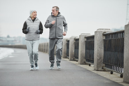 Positive senior Caucasian couple of joggers in warm clothes running along riverside with metal railings and talking to each otherの写真素材